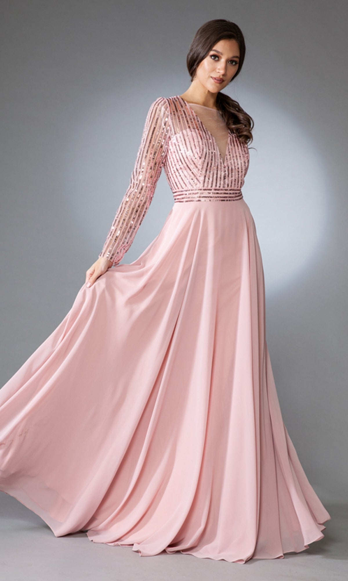 A woman stands against a plain gray background wearing the Long-Sleeve Long Modest Prom Dress 7036 in light pink, featuring a beaded bodice and flowing skirt.